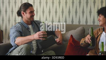 Lächelnde zwei Männer in legeren Kleidern sitzen zu Hause auf dem Sofa, halten grüne Bierflaschen und Tablets Stockfoto