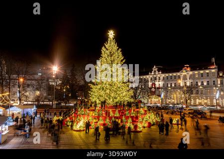 Ein hell erleuchteter Weihnachtsbaum auf einem festlichen Stadtplatz in Vilnius, Litauen, Europa, bei Nacht, umgeben von Menschen, Weihnachtsbeleuchtung und Marktstände Stockfoto
