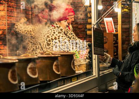 Ein Verkäufer serviert heißen Wein und heiße Schokolade an einem festlichen Weihnachtsmarkt in Vilnius, umgeben von dampfenden Töpfen und warmen Weihnachtslichtern Stockfoto