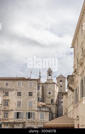 Ein sonnendurchflutetes europäisches Stadtbild mit verwitterten Steingebäuden, einer prominenten Uhr und einem Glockenturm sowie Dachterrassen mit Sonnenschirmen, die an alte Wo erinnern Stockfoto