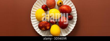 Eine Sammlung von bunten Ostereiern in verschiedenen Größen und Mustern wird auf einer weißen, gewellten Platte auf einer warmen braunen Oberfläche, b, ausgestellt Stockfoto