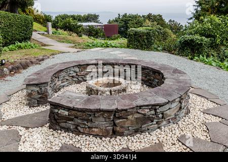 Eine kreisförmige Feuerstelle aus gestapelten Natursteinplatten in einem modernen Gartenpark mit Meerblick über die Sagami-Bucht in Odawara, Präfektur Kanagawa, Japan. Die Feuerstelle ist von einem Bett aus weißen Flusskieseln umgeben; ein gepflasterter Weg führt zu einem modernen Gebäude im Hintergrund Stockfoto