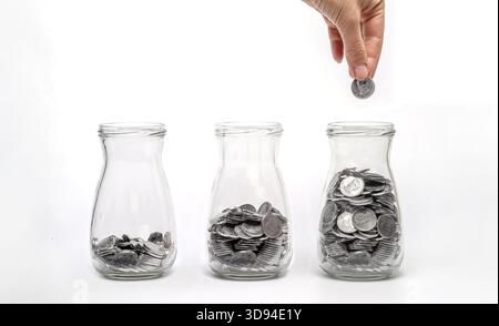 Drei Glasgefäße mit Münzen auf verschiedenen Ebenen, die von Hand mit Münzen gefüllt sind, symbolisieren das Finanzwachstum und die Einsparungen. Stockfoto