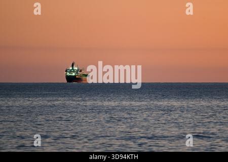 Ein beleuchtetes Frachtschiff segelt während Sonnenauf- oder -Untergang über ein ruhiges Meer. Der Himmel leuchtet in warmen Orange- und Rosa-Tönen, die sich auf dem Wasser spiegeln. Stockfoto