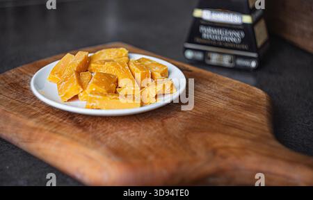 Aromatisch gereifter Käse, der auf einer Keramikplatte auf einem Holzbrett in feine Scheiben geschnitten wird, schafft eine raffinierte, handwerkliche Essensszene mit warmen Tönen und Stockfoto