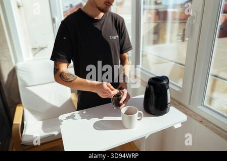 Junger Mann, der Instantkaffee in der Nähe des Fensters in hellem Arbeitsraum zubereitete Stockfoto
