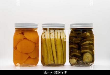 Drei mit Konserven gefüllte Gläser stehen auf einem einfachen weißen Hintergrund. Das linke Glas enthält Pfirsichscheiben, das mittlere Glas enthält eingelegte Gurken und das rechte Glas enthält Bananenscheiben in Sirup. Stockfoto