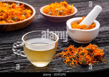 Calendula officinalis - Tasse Calendula-Tee, begleitet von getrockneten Blütenblättern und einem weißen Mörtel mit Calendula-Blüten Stockfoto