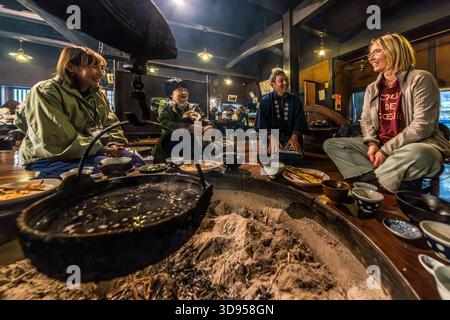 Vier Personen sitzen um ein Irori in einem traditionellen Restaurant in Hakone, Präfektur Kanagawa, Japan: ein Mitarbeiter in blauer Uniform und drei Gäste, darunter zwei nicht-japanische Frauen, unterhalten sich beim gemeinsamen Essen. Der Irori-Brazier im Amazake Chaya hängt von einem Haken mit einem hölzernen Wächterfisch, der vor Feuer schützen soll. Vor ihnen sind Keramikschalen mit japanischen Gerichten um die schwelende Feuerstelle angeordnet. Irori-Restaurants bieten den Gästen eine Einführung in die traditionelle Herd-Küche Japans Stockfoto
