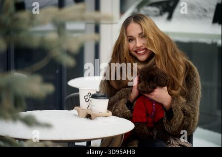 Modische junge Frau mit Pelzmantel, Schal und Sonnenbrille genießt Kaffee zum Mitnehmen in einem verschneiten Café im Freien, während sie einen niedlichen braunen Hund in der Hand hält Stockfoto