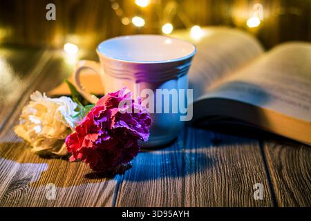 Kaffeetasse mit magentafarbener Nelke und weißer Blume vor offenem Buch und leuchtenden Lichtern, die Entspannung, Inspiration und kreatives Schreiben veranschaulichen Stockfoto