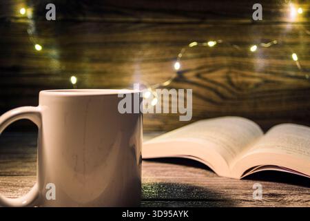 Minimales Stillleben mit einfarbigem, weißem Becher vor offenem Buch und warmen Lichterketten auf hölzernem Hintergrund, ideal für Achtsamkeit, Studium und Heim Stockfoto
