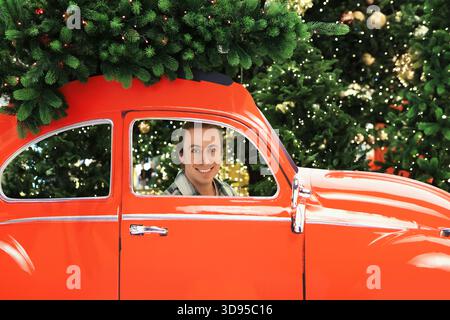 Glücklicher Mann, der in einem roten Retro-Auto sitzt, mit einem Weihnachtsbaum auf dem Dach vor einem festlichen Lichterhintergrund. Hochwertige Fotos Stockfoto