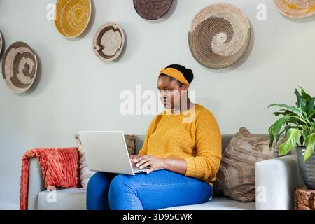 Afroamerikanerin mit Senfpullover, die auf dem Sofa sitzt und auf silbernem Laptop schreibt Stockfoto