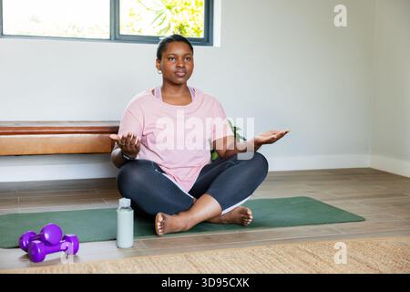 Afroamerikanerin meditiert auf grüner Matte in Yogaecke mit lila Kurzhanteln, Wasserflasche Stockfoto