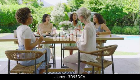 Eine Gruppe von fünf Frauen, die an einem rechteckigen Holztisch auf der Terrasse am Pool sitzen, mit Rosen Stockfoto