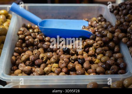Großpackung marinierter schwarzer und brauner Oliven mit blauer Schaufel, umgeben von grünen Oliven in Kunststoffbehältern Stockfoto