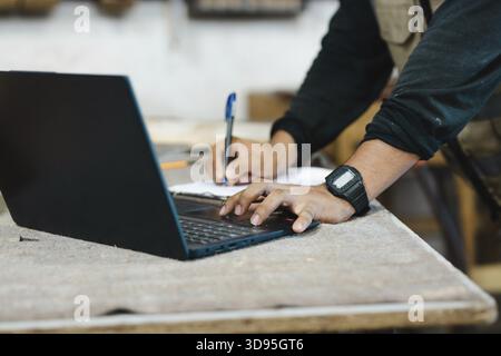 Nahaufnahme eines Zimmermanns mit einem Laptop an einer Werkbank. Nahaufnahme eines Mannes, der einen Laptop in der Tischlerei benutzt Stockfoto