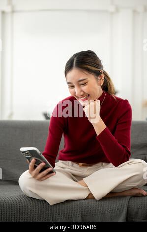 Eine hübsche asiatische Frau lächelte, als sie mit der Faust unter dem Kinn auf dem Sofa im Wohnzimmer oder in der Lounge saß. Zeit zur freien Verfügung zum Entspannen, Entspannen zu Hause. Stockfoto