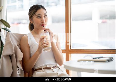 Eine hübsche asiatische Frau, die einen Strohhalm in einer Glasschale hält und Eiskaffee trinkt, während sie draußen vom Tisch des Cafés blickt. Zeit Zur Freien Verfügung Zum Entspannen, Entspannen Im Freien. Stockfoto