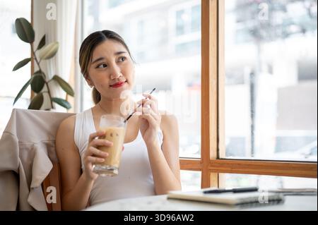 Hübsche asiatische Frau, die einen Strohhalm in einer Glasschale hält, nachdem sie Eiskaffee getrunken und sich vom Tisch des Cafés umgesehen hat. Zeit Zur Freien Verfügung Zum Entspannen, Entspannen Im Freien. Stockfoto