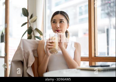 Hübsche asiatische Frau, die eine Glasschale hält und Eiskaffee mit Strohhalm trinkt, während sie sich vom Tisch des Cafés umsieht. Zeit Zur Freien Verfügung Zum Entspannen, Entspannen Im Freien. Stockfoto