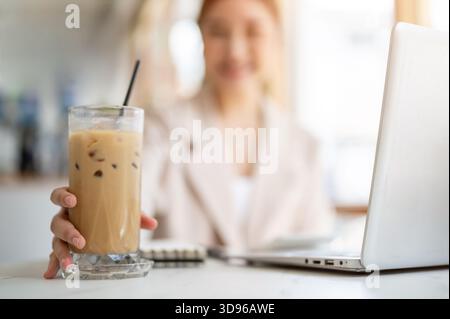 Nahaufnahme einer Frau mit der Hand, die Eiskaffee oder Milchtee auf einem Glasuntersetzer neben dem Laptop auf dem Tisch des Cafés hält oder schnappt. Arbeiten oder Lernen, Kühlen im Freien Stockfoto