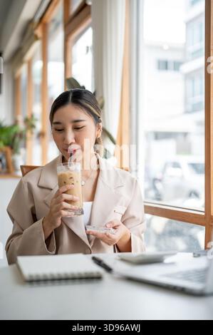 Hübsche asiatische Frau, die Glas coster hält, als Eiskaffee mit Strohhalm trinkt, während sie sich das Notizbuch auf dem Kaffeetisch ansieht. Arbeiten oder Lernen, Kühlen im Freien Stockfoto