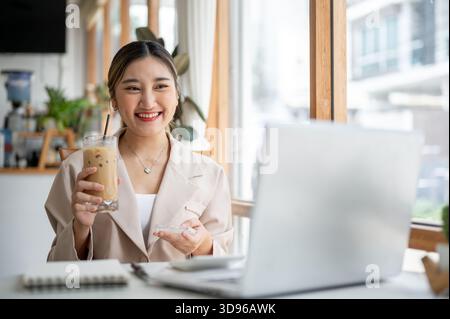 Eine hübsche asiatische Frau, die Eiskaffee mit einem Glas coster hält und auf den Laptop blickt, während sie am Tisch des Cafés sitzt. Arbeiten oder Lernen, Kühlen im Freien. Stockfoto