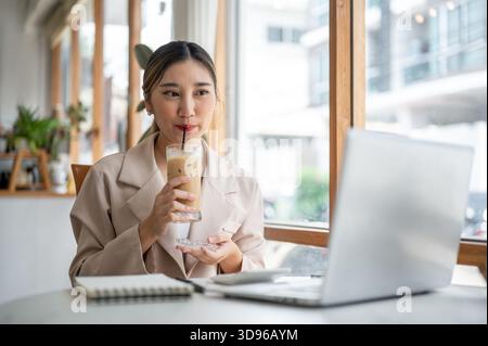 Hübsche asiatische Frau, die Glas coster hält, als Eiskaffee mit Strohhalm trinkt, während sie auf den Laptop auf dem Tisch des Cafés blickt. Arbeiten oder Lernen, Kühlen im Freien Stockfoto
