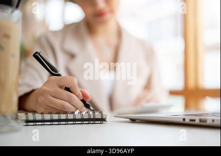 Eine asiatische Frau, die einen Füllfederhalter hält, der in Notizbuch schreibt, während sie mit Laptop und Kaffee am Kaffeetisch sitzt. Arbeiten oder Lernen, Kühlen im Freien Stockfoto
