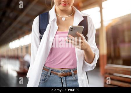 Nahaufnahme einer Frau mit Rucksack in der Hand und Blick auf das Smartphone beim Stehen oder Gehen am Bahnhof. Thailändische Eisenbahn, Fremdenverkehr. Stockfoto