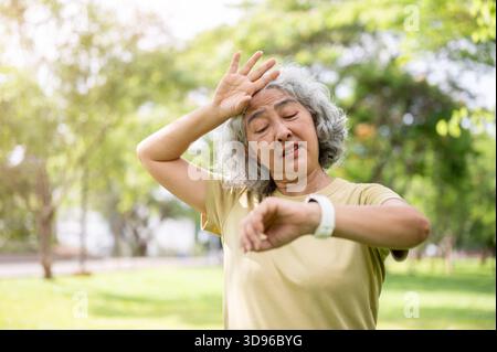 Alte Frau, die auf die Armbanduhr schaut, während sie sich nach dem letzten Training im Park ihren Schweiß abwischt. Älteres Ehepaar, grüne Natur, chilliger Outdoor. Stockfoto