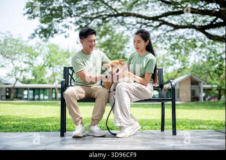 Lächelnder asiatischer Mann und eine asiatische Frau, die einen Corgi-Welpen halten und klopfen und sich ausruhen, nachdem sie auf der Parkbank sitzen. Ein paar Hemd, ein Hund, ein Familienausflug. Stockfoto