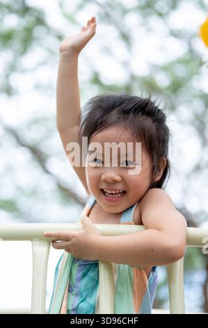Lächelndes asiatisches Mädchen, das auf dem Spielplatz des Parks auf einer bunten Kletterrutsche die Hand erhebt. Kreative Spielsachen, süße Kinder, Familienausflug. Stockfoto