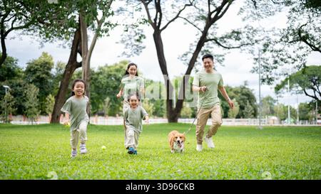 Dad hält eine Leine an Corgi-Welpen, während er mit Mutter Sohn und Tochter im Park läuft oder läuft. Eltern und Kinder, Hund, Familienausflug. Stockfoto