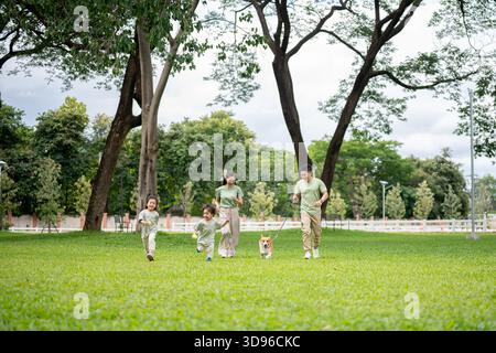 Dad hält die Leine an einem Corgi-Welpen, während er mit Mutter und Tochter auf Gras im Park läuft. Eltern und Kinder, Hund, Familienausflug. Stockfoto