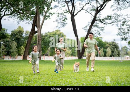 Dad hält eine Leine an Corgi Welpen, während er läuft, und Mutter mit Sohn und Tochter auf Gras im Park. Eltern und Kinder, Hund, Familienausflug. Stockfoto