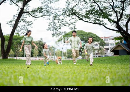 Lächelnder Vater hält eine Leine an einem Corgi-Welpen, während er mit Mutter Sohn und Tochter auf dem Gras im Park läuft. Eltern und Kinder, Hund, Familienausflug. Stockfoto