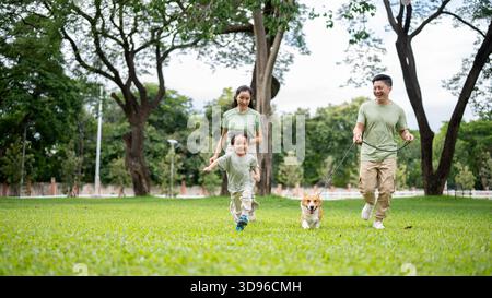 Lächelnder Vater hält eine Leine am Corgi-Welpen, während er spaziert oder läuft, Mama und Sohn auf Gras im Park. Eltern und Kinder, Hund, Familienausflug. Stockfoto