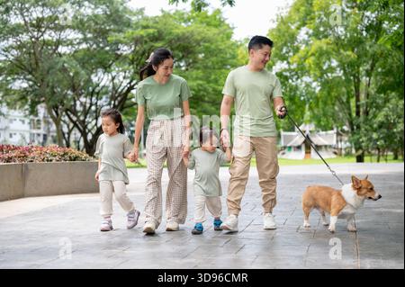 Lächelnder Vater und Mutter mit Sohn und Tochter, die eine Leine am Corgi-Welpen halten, während sie im Hof im Park spazieren. Eltern und Kinder, Hund, Familienausflug. Stockfoto