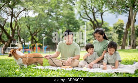 Dad hält Corgi-Welpen-Leine als Mutter mit Sohn-Tochter, die auf Picknickmatten sitzt und auf Gras im Park liegt. Eltern und Kinder, Hund, Familienausflug Stockfoto