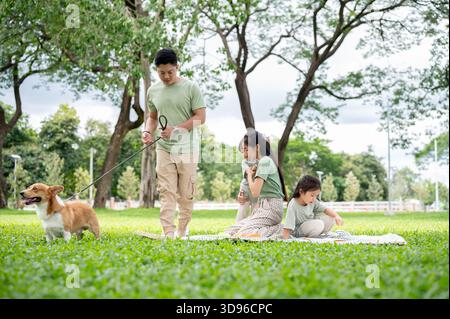 Dad hält die Leine an einem Corgi-Welpen, und Mom hält Sohn und Tochter, die auf Picknickmatten über dem Gras im Park sitzen. Eltern und Kinder, Hund, Familienausflug. Stockfoto