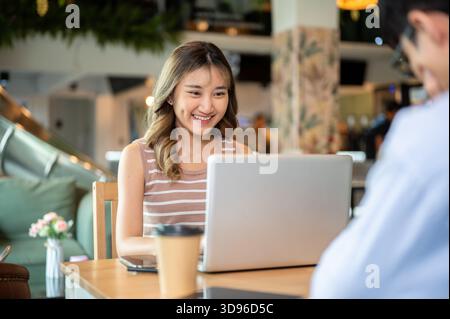 Asiatische Frau, die mit einem Mann am Holztisch im Café oder in der Kantine sitzt und auf den Laptop blickt. In Arbeit oder Studie, Aufgabe oder Gruppenprojekt. Stockfoto