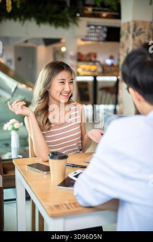 Asiatische Frau lächelt und spricht einem Freund, während sie am Holztisch im Café oder Kantine sitzt. In Arbeit oder Studie, Aufgabe oder Gruppenprojekt Stockfoto