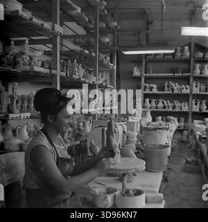 Slawjansk, Ukraine, UdSSR. 1968. Schwarzweiß-Archivfoto, das eine Modelliererin des Keramikkomplexes Slavjansk zeigt, wie sie eine Tonfigur von Hand modelliert. Der Arbeiter, der ein ärmelloses Kleid und eine stilvolle Bienenstockfrisur aus den 1960er Jahren trägt, sitzt an einem Tisch mit Schüsseln aus Ton. Holzregale im Hintergrund sind mit trockenem rohen Keramikstatuen, darunter Vögel und menschliche Figuren, gefüllt. Dieses industrielle Image fängt die Herstellung von Porzellan-Konsumgütern im friedlichen Donbass vor dem Krieg ein. Stockfoto