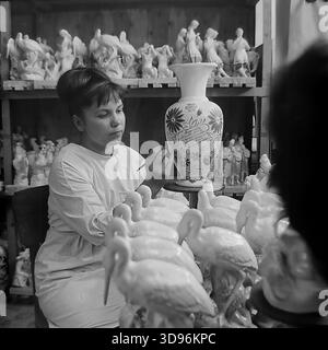 Slawjansk, Ukraine, UdSSR. 1968. Schwarzweiß-Archivfoto einer Künstlerin im Slavjansk Ceramic Combine Gemälde eine große Gedenkvase, die dem 50. Jahrestag des Komsomols (VLKSM) gewidmet ist. Die Frau sitzt in einer Werkstatt, umgeben von Regalen mit keramischen Konsumgütern, darunter Figuren von Störchen und Mädchen in Nationalkostümen. Das Bild fängt die zarte Handarbeit und Massenproduktion von Porzellandekor im friedlichen Donbass vor dem Krieg ein. Stockfoto