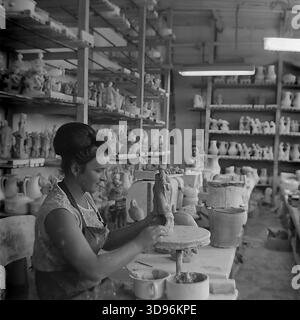 Slawjansk, Ukraine, UdSSR. 1968. Schwarzweiß-Archivfoto, das eine Modelliererin des Keramikkomplexes Slavjansk zeigt, wie sie eine Tonfigur von Hand modelliert. Der Arbeiter, der ein ärmelloses Kleid und eine stilvolle Bienenstockfrisur aus den 1960er Jahren trägt, sitzt an einem Tisch mit Schüsseln aus Ton. Holzregale im Hintergrund sind mit trockenem rohen Keramikstatuen, darunter Vögel und menschliche Figuren, gefüllt. Dieses industrielle Image fängt die Herstellung von Porzellan-Konsumgütern im friedlichen Donbass vor dem Krieg ein. Stockfoto