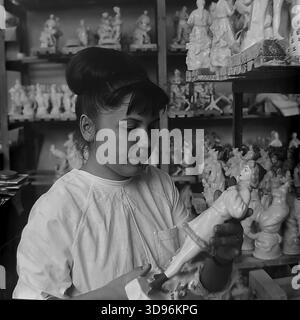 Slawjansk, Ukraine, UdSSR. 1968. Schwarzweiß-Archivfoto, auf dem eine Angestellte des Keramikkomplexes Slavjansk die Qualitätskontrolle von Porzellanerzeugnissen durchführt. Die Frau im weißen Arbeitsmantel inspiziert eine kleine Figur in ihren Händen. Hinter ihr befinden sich Holzregale mit Reihen identischer Statuetten, die Mädchen in Wintermänteln und -Muffen darstellen. Das Bild zeigt die Massenproduktion von Souvenirs und die Liebe zum Detail im friedlichen Donbass vor dem Krieg. Stockfoto