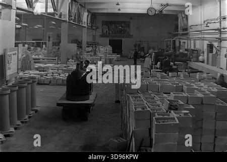 Slawjansk, Ukraine, UdSSR. 1968. Schwarzweiß-Archivfoto, das das Innere eines Lagerhauses im Keramikkompanie Slavjansk zeigt. Ein männlicher Arbeiter zieht einen Wagen mit großen Hochspannungs-Porzellan-Isolatoren. Der Vordergrund ist gefüllt mit Reihen von Keramikrohren und gestapelten Holzkisten mit Produkten. Im Hintergrund ruft ein sowjetisches Propagandaplakat an der Wand dazu auf, Strom zu sparen. Das Bild zeigt das Ausmaß der industriellen Produktion im friedlichen Donbass vor dem Krieg. Stockfoto